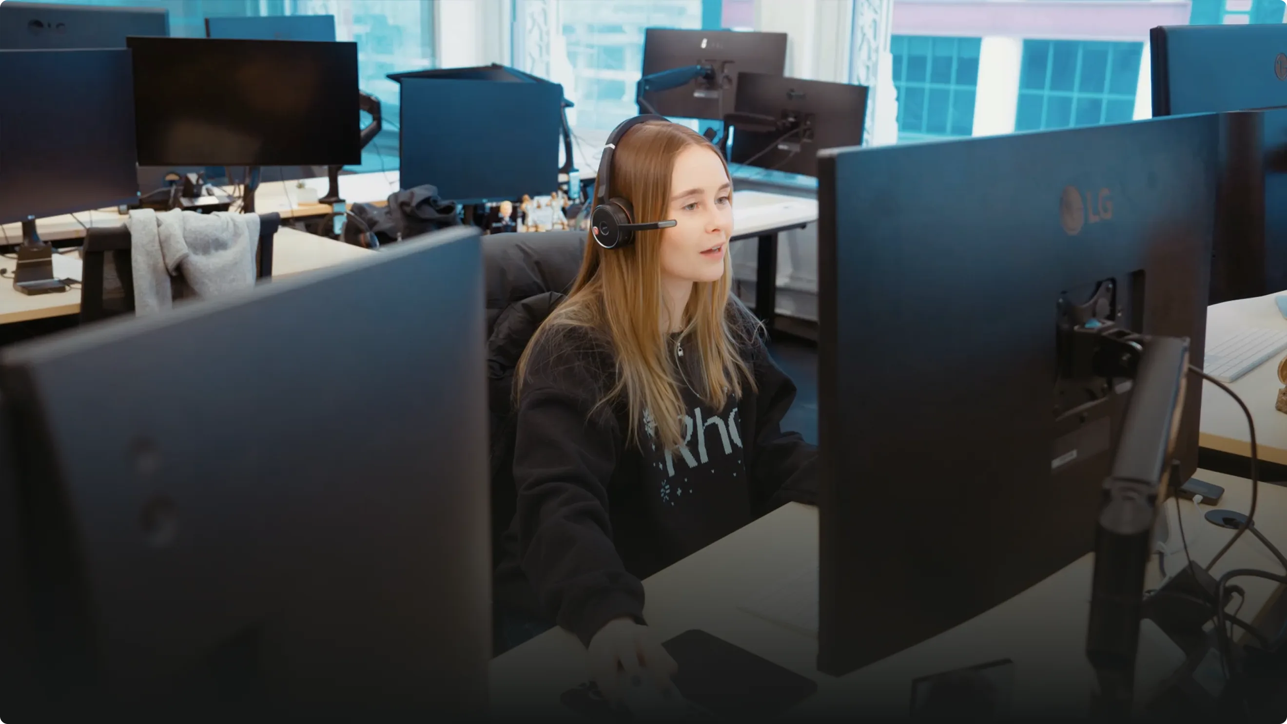 A woman wearing a headset works at a computer in a modern office with multiple monitors and large windows.